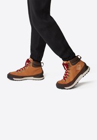 Botas de senderismo marrones con cordones rojos, material de ante y cuero, diseño de caña alta, suela de goma y ojetes de metal para los cordones.