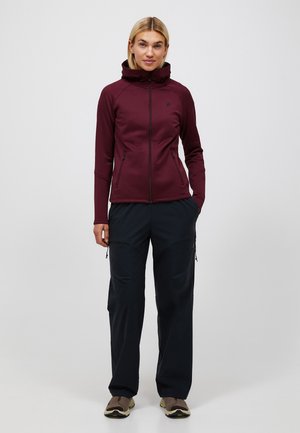 Kvinde stående iført en bordeauxfarvet lynlåsjakke, sorte cargobukser og brune sneakers mod en ensartet lys baggrund.