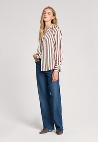 Pasiasta koszula z długim rękawem w kolorze kremowym i burgundowym, zestawiona z szerokimi niebieskimi dżinsami. Charakteryzuje się luźnym krojem.