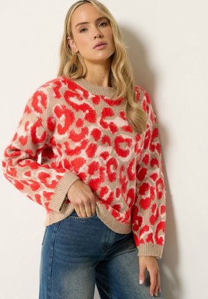 Femme aux longs cheveux blonds portant un pull beige à imprimé léopard rouge et un jean bleu, debout contre un mur blanc.
