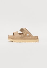 Beige suède slidesandaal met twee verstelbare banden, circulaire logo-hardware en een gestructureerde zool, met een ronde neus en minimalistisch ontwerp.