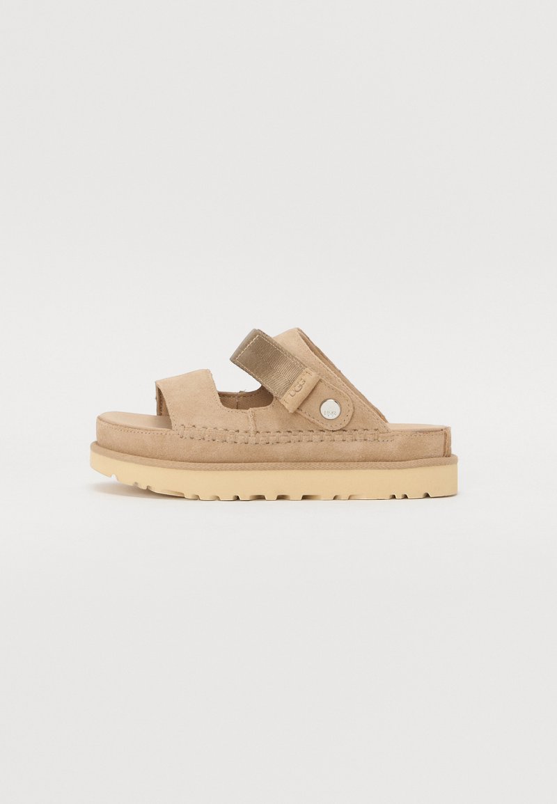Beige suède slidesandaal met twee verstelbare banden, circulaire logo-hardware en een gestructureerde zool, met een ronde neus en minimalistisch ontwerp.