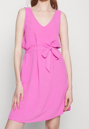 Kobieta w różowej sukience bez rękawów z dekoltem w serek i wiązanym paskiem w talii, stojąca na tle jednolitego jasnego tła.