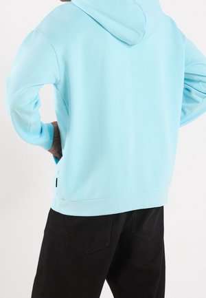 Persoon die een lichtblauwe hoodie draagt met de capuchon naar beneden en een zwarte broek, met de handen in de zij staand, de rug naar voren gericht.