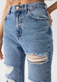 Jeans de mezclilla azul claro con cintura alta, que presentan desgarros en las rodillas, bordes deshilachados y herrajes de cobre para el cierre.