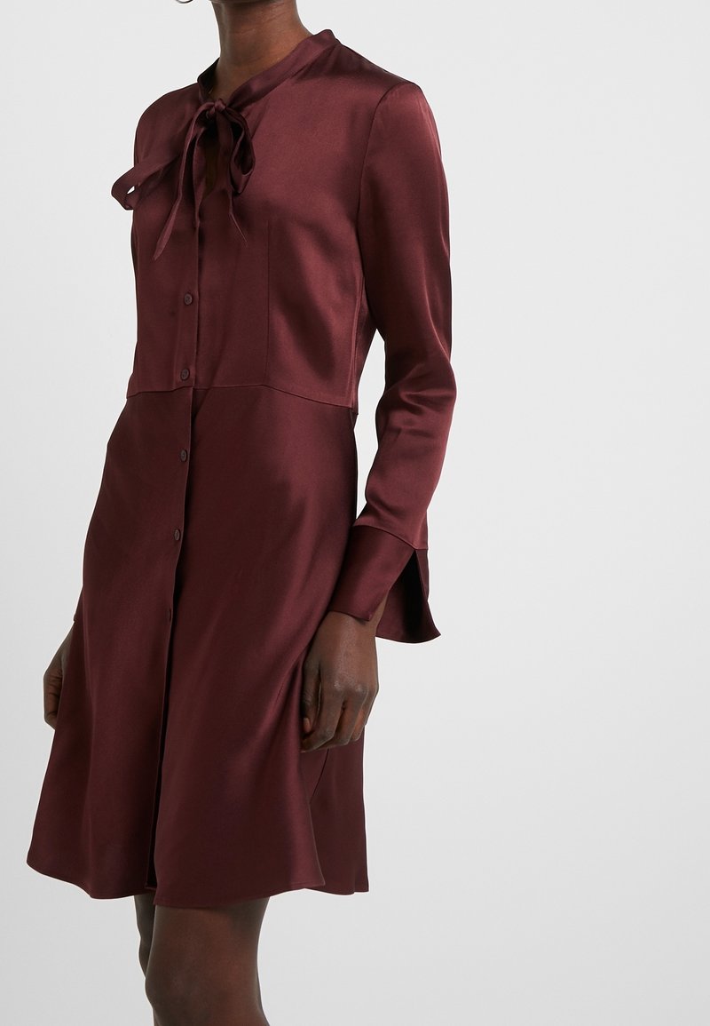Femme portant une robe bordeaux à manches longues, boutonnée devant, avec un nœud noué au col et une jupe évasée.