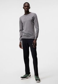 Pilkas megztinis su apvalia iškirpte, derinamas su juodomis kelnėmis ir sportbačiais, kuriuose yra žalių akcentų, stovintis priešais vienalytį foną.