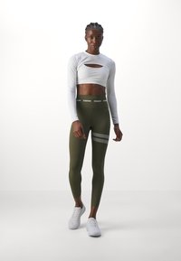 Haut court à manches longues blanc avec un décolleté découpé, associé à des leggings vert olive avec un détail à rayures grises et une taille "STRONGER".