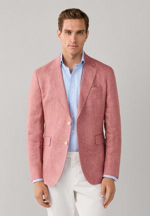 Man draagt een roze blazer over een lichtblauw overhemd en witte broek tegen een effen grijze achtergrond.