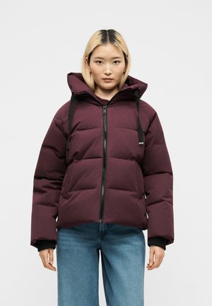 Junge Frau mit glatten blonden Haaren, die eine bordeauxrote gesteppte Jacke mit Reißverschluss und blaue Jeans trägt, steht vor einem schlichten weißen Hintergrund.
