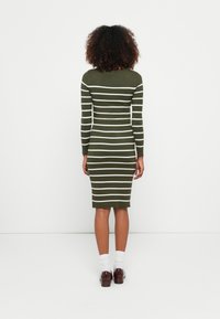 Donna con capelli ricci che indossa un vestito aderente verde oliva con righe orizzontali bianche, calze bianche e scarpe marroni, in piedi di fronte a una parete bianca.