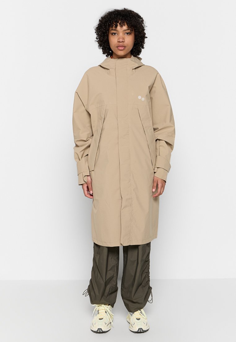 Beige wasserfester Trenchcoat mit hohem Kragen und Fronttaschen. Mit lockeren Ärmeln und zwei runden Akzenten nahe der Brust.