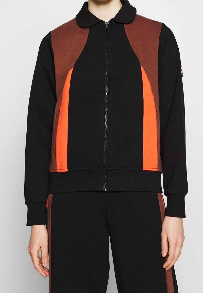 Personne portant une veste noire zippée avec des panneaux marron et orange, associée à un pantalon noir assorti avec des bandes latérales marron.
