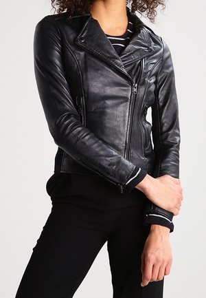 Persoon draagt een nauwsluitend zwart leren motorjack over een zwart-wit gestreept topje, met een zwarte broek en armen gekruist ter hoogte van de taille.