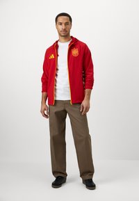 Giacca rossa con zip e dettagli gialli, con emblema della Spagna; indossata sopra una maglietta bianca. I pantaloni marroni e le sneaker nere completano l'outfit.