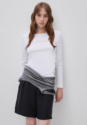Jonge vrouw met lang bruin haar, die een wit shirt met lange mouwen met tekst draagt en een zwarte korte broek, een grijze trui om haar middel geknoopt.
