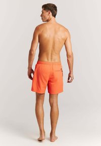 Modello maschile in piedi a piedi nudi, visto di spalle, indossa pantaloncini da bagno arancioni vivaci con una tasca posteriore, su uno sfondo chiaro e semplice.