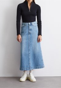 Frau in einem schwarzen gerippten Langarmoberteil, hellblauem Denim-Midirock, weißen Socken und weißen Sneakern, die an einer weißen Wand steht.