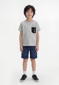 Băiat stând cu o expresie neutră, purtând un tricou gri cu un imprimeu negru pe buzunar, pantaloni scurți din denim, șosete albe și adidași negri.