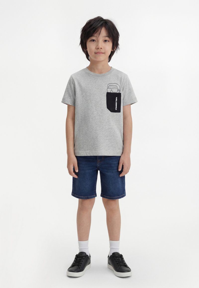 Băiat stând cu o expresie neutră, purtând un tricou gri cu un imprimeu negru pe buzunar, pantaloni scurți din denim, șosete albe și adidași negri.