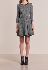 Robe en tweed gris avec un buste ajusté, une jupe évasée, des manches trois quarts et un accent noir au niveau du col. Portée avec des bottines noires.