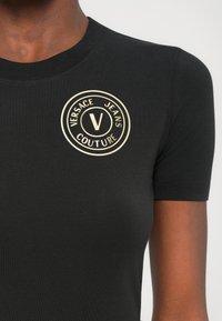 Melna īsām piedurknēm T-kreklu ar rievotu tekstūru, kas rotāta ar apaļu zelta logo, kurā rakstīts "VERSACE JEANS COUTURE", centrā uz krūšu daļas.