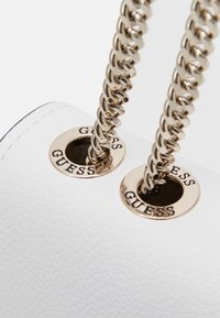 Coin d'un sac à main blanc texturé avec deux poignées en chaîne dorée passant à travers des œillets dorés gravés du nom de la marque "GUESS".