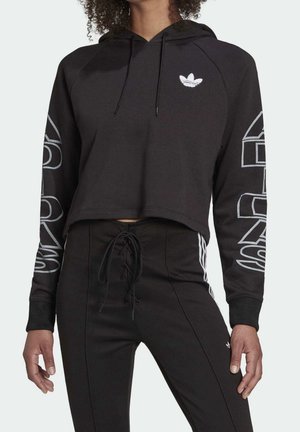 Osoba nosząca czarną skróconą bluzę z kapturem Adidas z logo na klatce piersiowej i napisem na rękawach, w połączeniu z czarnymi spodniami z wysokim stanem i białymi paskami.