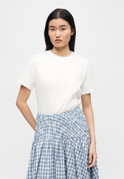 Νεαρή γυναίκα με μακριά μαύρα μαλλιά που φορά απλό λευκό κοντομάνικο μπλουζάκι και φούστα με μπλε και λευκό μοτίβο σε απλό φόντο.