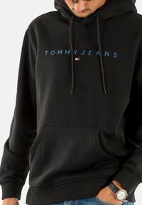 Felpa nera con testo blu "TOMMY JEANS". Presenta una tasca frontale, cordini e polsini a coste. Realizzata in un tessuto morbido e strutturato.