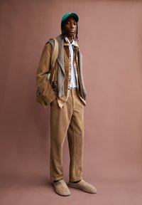 Tan corduroy jas en broek met een grijze sweatshirt over de schouders. Lichtblauw shirt daaronder. Groene pet en beige instapschoenen.