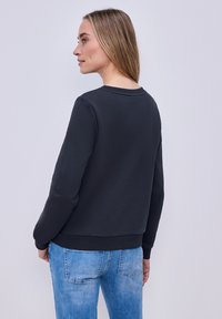 Femme aux cheveux châtain clair portant un sweat-shirt noir à manches longues et un jean bleu, debout de profil sur un fond clair uni.