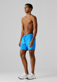 Noor mees seisab paljalt, seljas erk sinised ujumispüksid ja valged tennisekingad, taustal hall ühtlane taust.