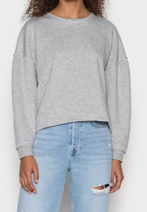 Person wearing light gray long-sleeve sweatshirt with raw seam details paired with light blue ripped jeans, standing against a plain background.