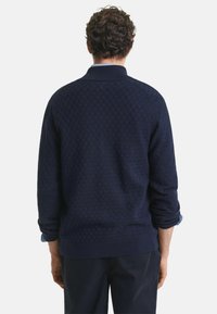 Suéter azul marino con patrón de cuadros texturizado, cuello y puños acanalados, corte relajado, llevado sobre una camisa azul clara, vista trasera.