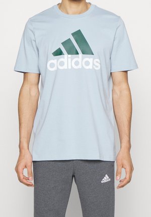Man die een lichtblauw Adidas T-shirt met donker groen logo en donkergrijze Adidas trainingsbroek draagt, staand tegen een effen achtergrond.