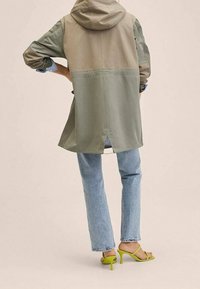 Veste légère bicolore avec capuche ; blocs de couleur vert olive et beige ; coupe décontractée avec une fente à l'arrière ; associée à un jean bleu clair et des talons vert vif.