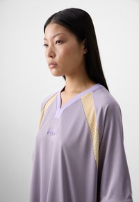 Giovane donna con lunghi capelli lisci, che indossa un top sportivo lavanda con dettagli in rete. Ha un'espressione neutra su uno sfondo chiaro.