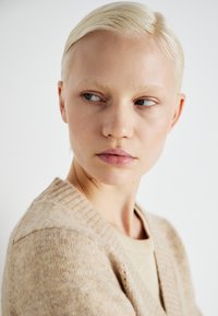 Beige κασκόλ με ραμμένο γιακά, μαλακή υφή και ανοιχτό μπροστά. Το μοντέλο έχει κοντά, καλοχτενισμένα ξανθά μαλλιά και διακριτικό μακιγιάζ.
