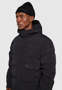 Sort puffer-jakke med hætte, kviltet design, lomme med lynlås fortil og brandingdetalje. Accepteret af en sort strikket hue.