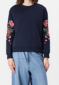 Sudadera marina con bordado floral en tonos rosa y rojo en ambas mangas, combinada con jeans de color azul claro.