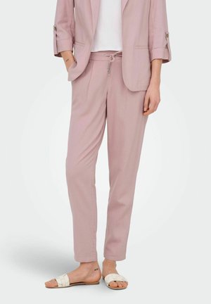 Kvinde iført lyserøde skræddersyede bukser med snøre i taljen, matchende blazer, hvid top og hvide slip-in sandaler med flettede remme.