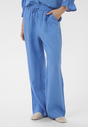 Nő laza, világoskék lenvászon nadrágot visel, elasztikus derékrésszel és húzózsinórral, fehér, szőtt, könnyen felvehető cipővel párosítva, zsebébe dugott kézzel áll.