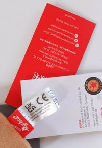 Red product tag with white text, featuring contact information and symbols. Includes a gray fabric label with CE markings and a logo.