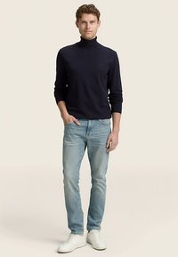 Suéter de cuello alto azul marino de tela suave, combinado con jeans azul claro y zapatillas blancas. Modelo de pie, erguido, contra un fondo neutro.