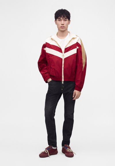 Jeune homme portant une veste zippée rouge et beige, un jean noir et des baskets rouges, debout devant un fond blanc uni.