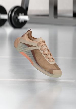 Baskets athlétiques marron clair et beige avec semelle translucide flottant au-dessus du sol de la salle de sport, poids flous en arrière-plan.