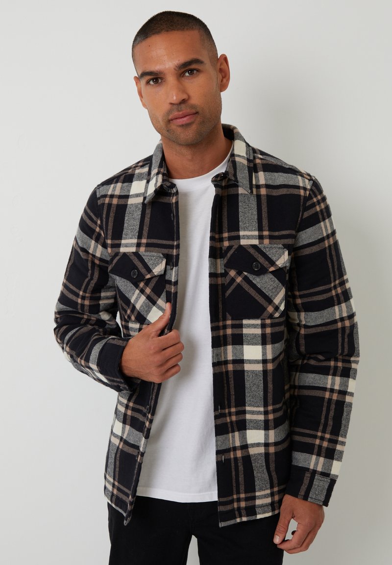 Camicia in flanella a quadri neri e grigi con accenti crema e marrone, dotata di due tasche sul petto e chiusura a bottoni. Indossata sopra una camicia bianca.