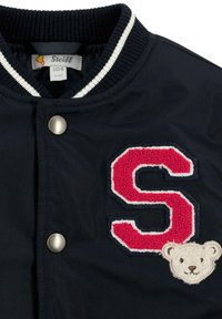 Giubbotto bomber navy con colletto a costine, caratterizzato da una "S" rossa ricamata e una toppa di un orso. Include bottoni a pressione argentati.