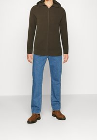 Olivgrön zip-up hoodie med en texturerad stickad design, med en huva, bärs över blå denimjeans och bruna stövlar.